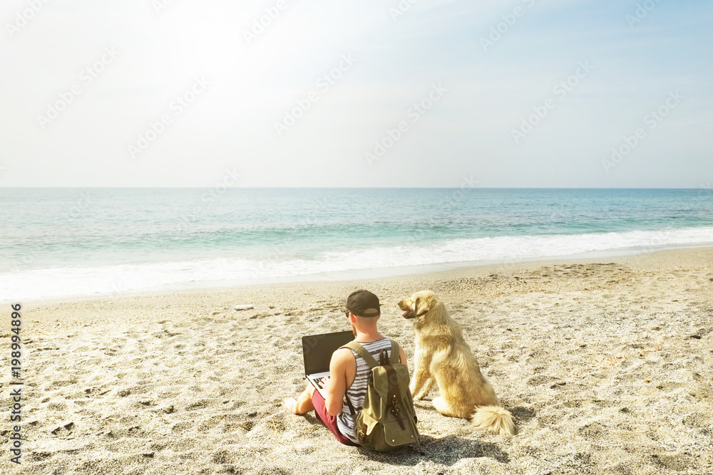 Rear view of man & his dog sitting at beach watching ocean waves, clear sunny day. Fit programmer freelancer male working on laptop computer at sea w shepherd canine. Background, copy space, landscape