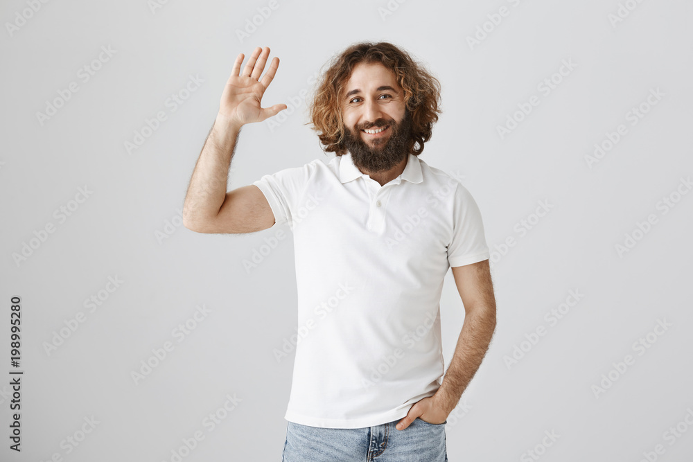 Fototapeta premium Hi, my friend. Portrait of good-looking ordinary eastern male with curly hair and beard raising hand and waving in greeting gesture, welcoming familiar person on street or giving five to mate