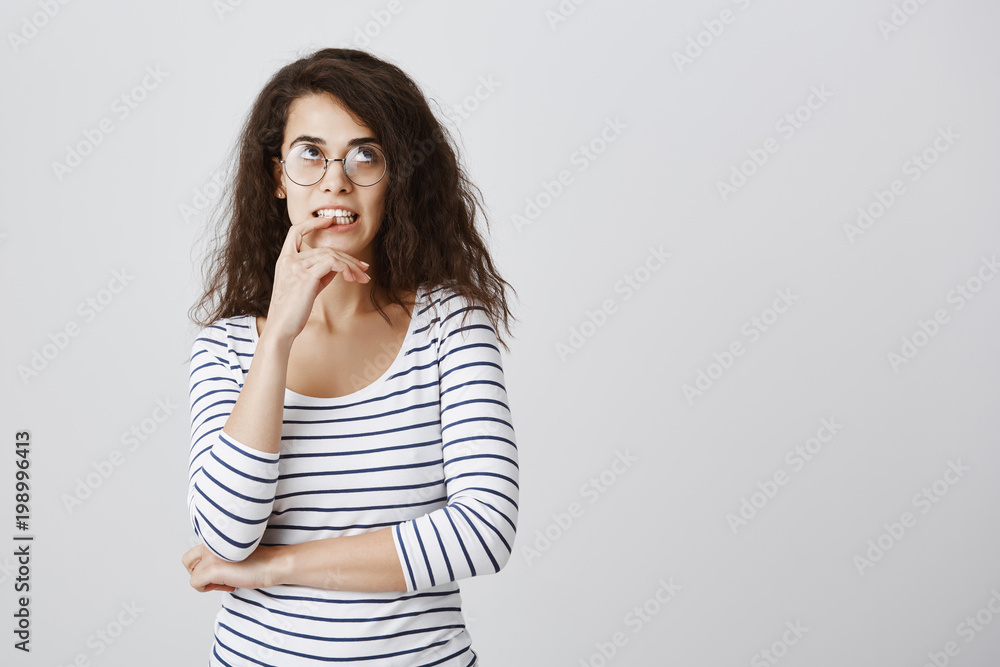 Attractive nerdy girl trying to calculate in mind. Portrait of focused entertained cute curly-haired female in glasses looking up, touching teeth with finger while thinking or trying to solve riddle Attractive nerdy girl trying to calculate in mind. Portrait of focused entertained cute curly-haired female in glasses looking up, touching teeth with finger while thinking or trying to solve riddle