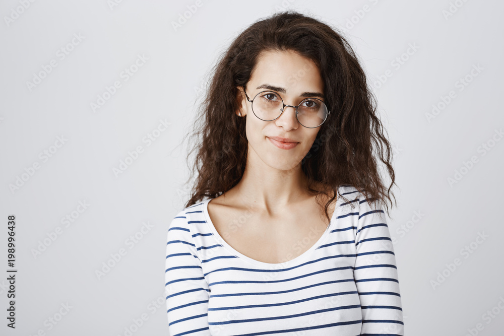 Waist-up shot of cute smart curly-haired girl in round glasses smiling broadly, being able to solve any riddle or issue, standing confident against gray background. Freelancer having break on coffee