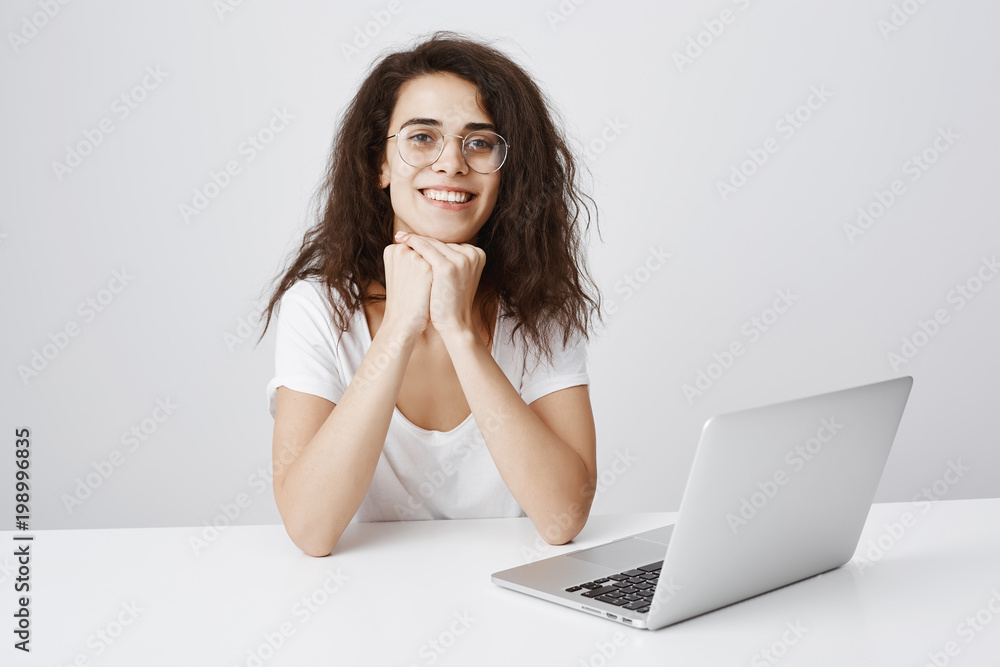Keep talking, I am interested. Studio shot of smiling curious curly-haired female leaning on hands and listening intriguing story, sitting at table near laptop, involved in conversation Keep talking, I am interested. Studio shot of smiling curious curly-haired female leaning on hands and listening intriguing story, sitting at table near laptop, involved in conversation