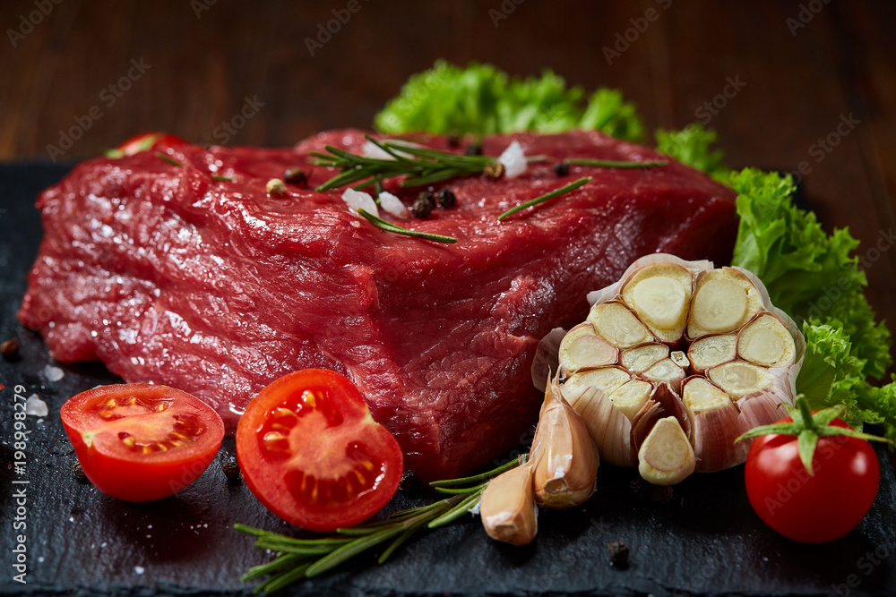 Composition of raw beefsteak on slate board with vegetables and seasoning, selective focus, close-up.