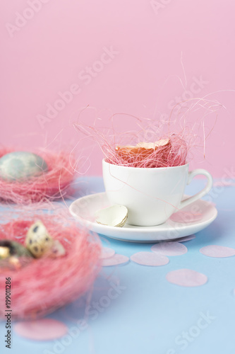 Yolk of broken egg in shell in a white coffee cup on a saucer, and blue chicken and quail eggs decorated with a pink sisal and confetti on a blue and pink background