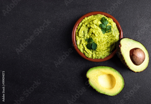 Guacamole sauce  in clay bowl with  cut half avocado on dark background. Top view with copy space.