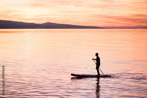 Silhouette of man at sunset standing on paddle board. Summer beach leisure activity. Active healthy lifestyle and travel summer concept
