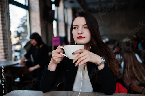 Brunette girl sitting on cafe with cup of cappuccino  listening music on headphones.