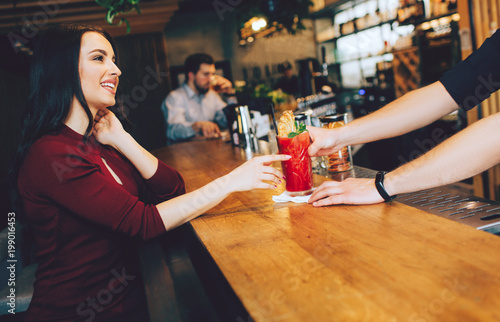 Attractive and gorgeous woman sitting at the stand and getting her order from barman. It is red cocktail. She looks to barman and smiling. Also there is another customer sitting not very far from her. photo