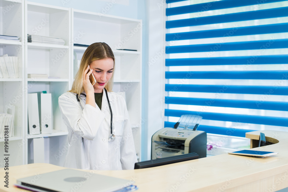 Portrait of a beautiful smiling nurse at desk station while talking on the phone and complete a medical information form Portrait of a beautiful smiling nurse at desk station while talking on the phone and complete a medical information form