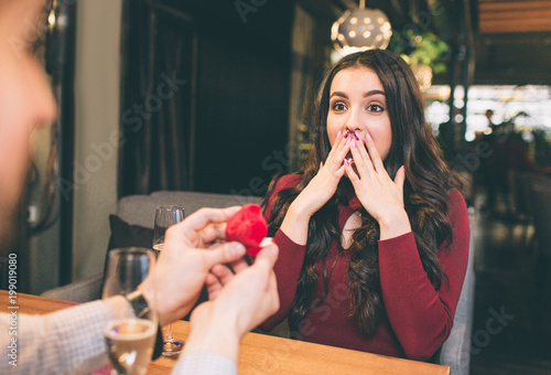 Good shot of young man doing propose to his beloved woman. He is showing her a box with ring inside it. She looks amazed and surprised. She didn't expect that. photo