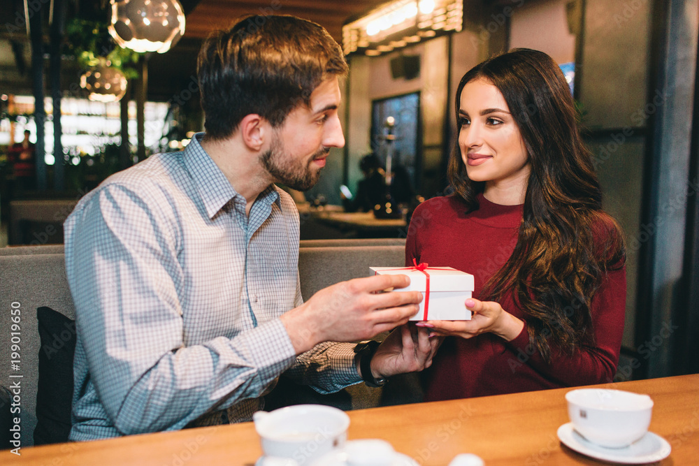 Picture of man giving a present to a woman. They are looking to each other and smiling a bit. They are sittin in restaurant.