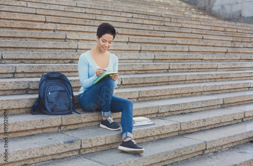 Pretty shorthaired woman sitting on the steps writing in notebook outdoors © Prostock-studio