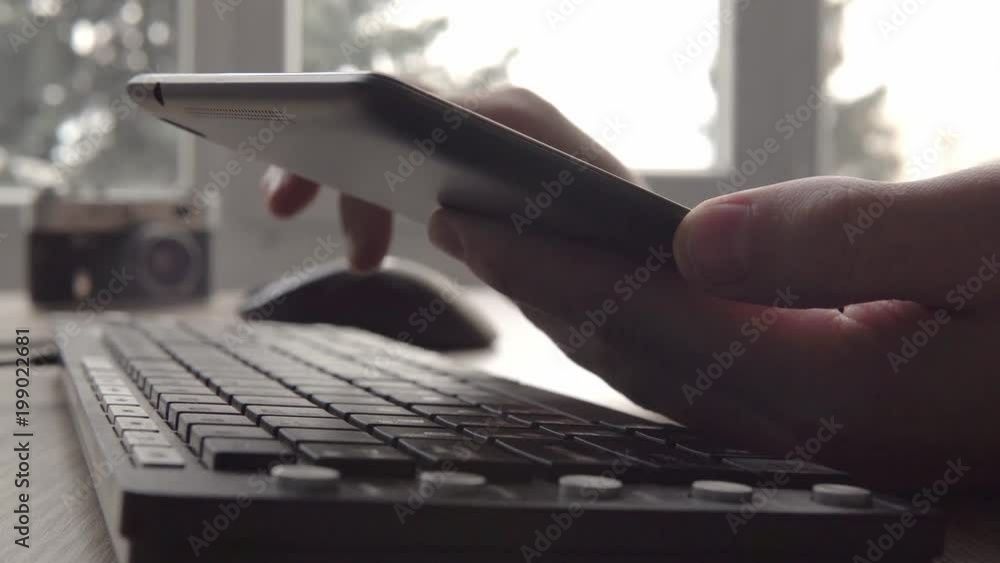 Close up on hands of young man using tablet while sitting computer keyboard and mouse. Freelancer photographer working and using tablet for communication with clients.