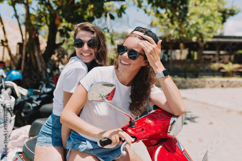 Two young beautiful hipster women riding on motorbike, summer island vacation, traveling, smiling, happy, having fun, sunglasses, stylish outfit, adventures, positive, friends together © PhotoBook