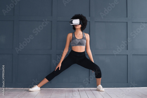 Sport in virtual reality! A graceful beautiful young woman practicing sports exercise on the grey background and posing for camera in 3D glasses.