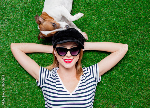 Redhead girl in sunglasses and hat with dog lying down on green spring time grass. © Masson