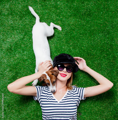 Redhead girl in sunglasses and hat with dog lying down on green spring time grass. © Masson