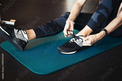 Young attractive brunette girl enlacing her sport shoes after practicing workout and crossfit training on blue yoga mat. Blurred background and focus on young woman side view.