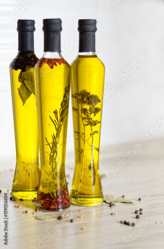 Three bottles of olive oil with spices, rosemary, pepper, oregano, allspice, bay leaf and pot with rosemary on the kitchen table, colorful pepper seeds in the foreground. Selective focus.