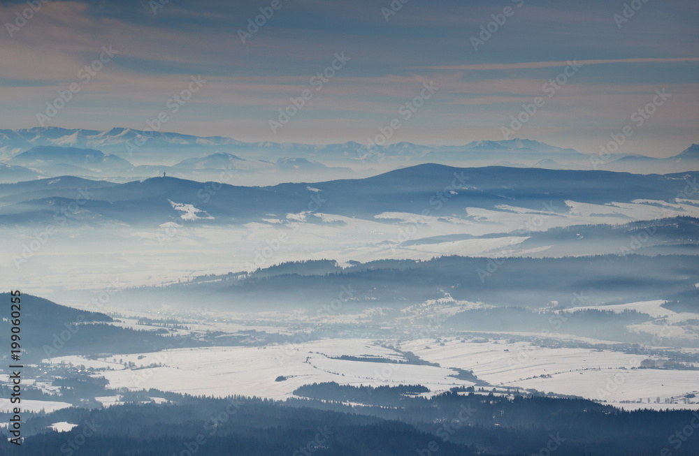 Sunlit winter scenery elevated view with blue mountain ridges, snowfields in misty valleys and Low Tatras in the distance, Orava and Liptov regions Oravska Magura Chocske vrchy ranges Slovakia Europe