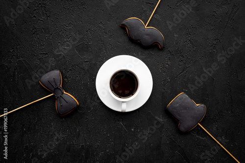 Father's Day celebration concept. Monochrome. Cookies in shape of moustache, hat, bow tie near coffee on black background top view