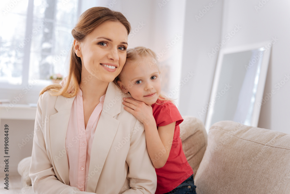 Happy family. Nice cute blonde girl sitting together with her mother and smiling while looking at you Happy family. Nice cute blonde girl sitting together with her mother and smiling while looking at you