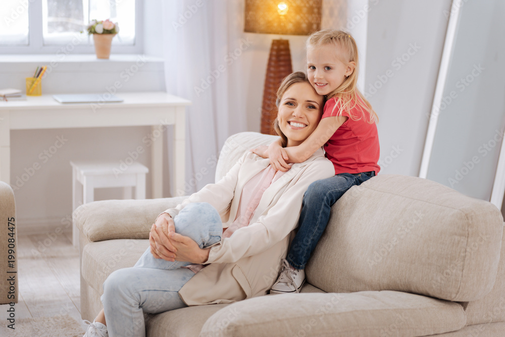 Family love. Cute nice smiley girl sitting on the sofa and hugging her mother while being at home together with her