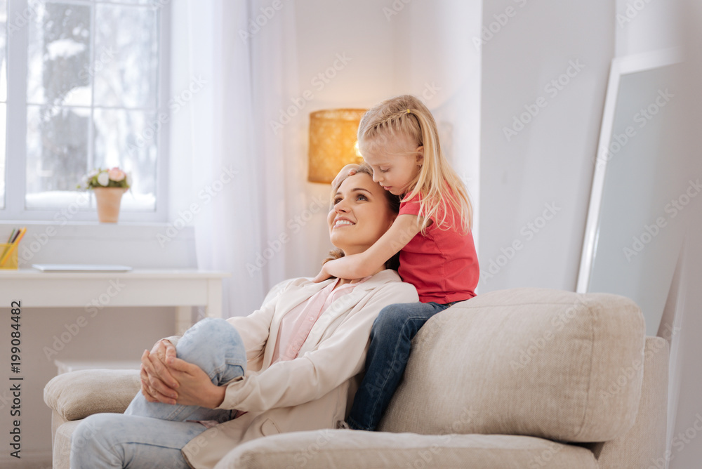 Positive emotions. Joyful nice positive mother sitting on the sofa and smiling while being hugged by her daughter Positive emotions. Joyful nice positive mother sitting on the sofa and smiling while being hugged by her daughter
