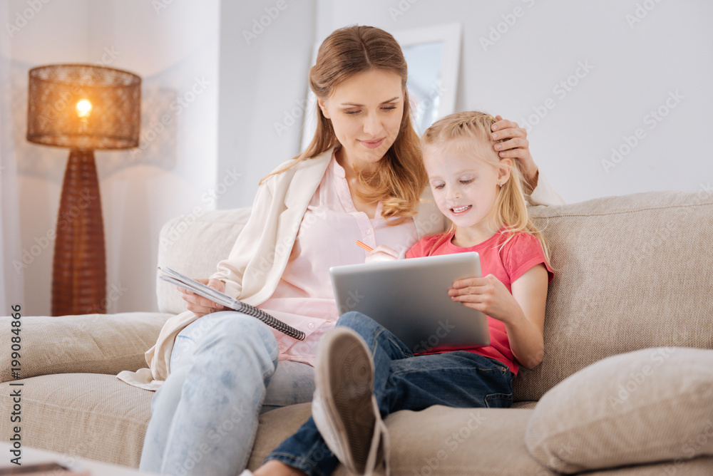Developing app. Nice smart positive girl sitting together with her mother and using a tablet while playing a developing game Developing app. Nice smart positive girl sitting together with her mother and using a tablet while playing a developing game