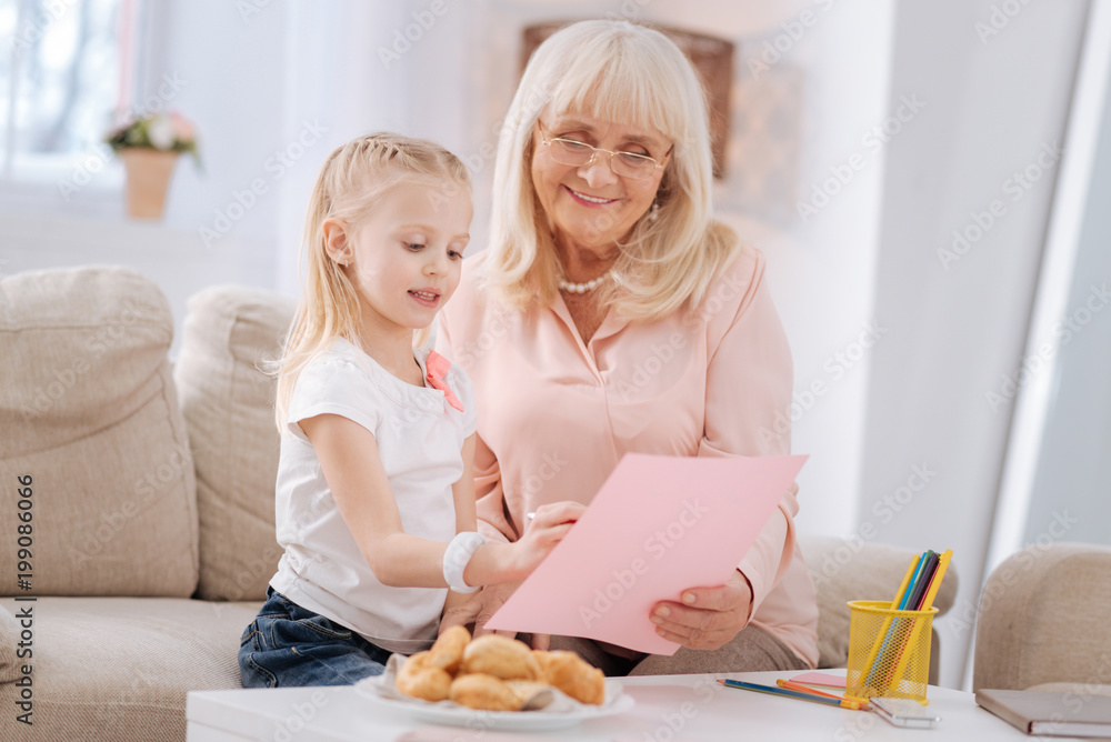 Talented child. Cheerful positive aged woman holding a drawing and smiling while talking to her granddaughter Talented child. Cheerful positive aged woman holding a drawing and smiling while talking to her granddaughter