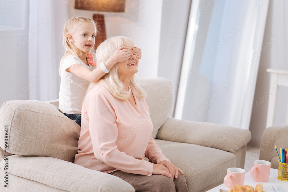 Guess who. Pleasant little girl closing the eyes of her granny at home Guess who. Pleasant little girl closing the eyes of her granny at home