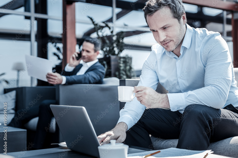 Modern device. Delighted positive nice businessman smiling and looking at the laptop screen while working on it Modern device. Delighted positive nice businessman smiling and looking at the laptop screen while working on it