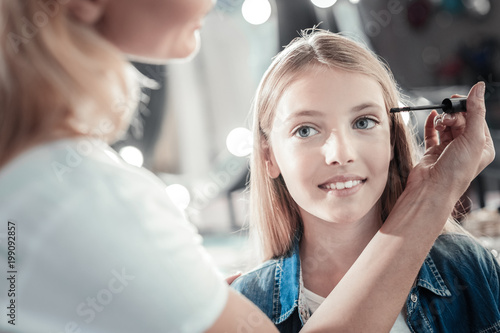 Great mood. Cheerful positive cure girl smiling and looking in front of her while having her eyelashes painted