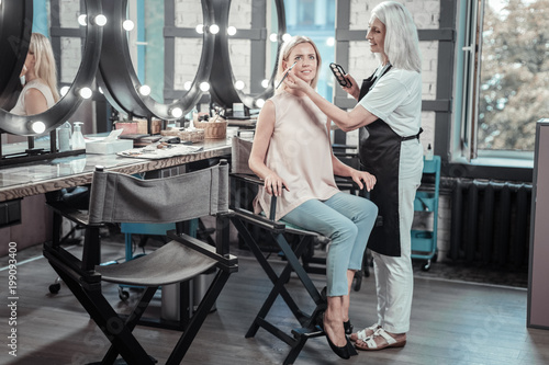 Professional salon. Delighted positive nice woman sitting on the chair and smiling while having her makeup done