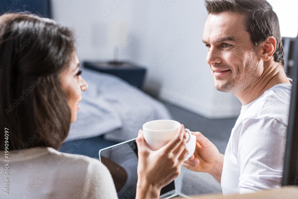 multiethnic couple holding cups of coffee and smiling each other at home multiethnic couple holding cups of coffee and smiling each other at home