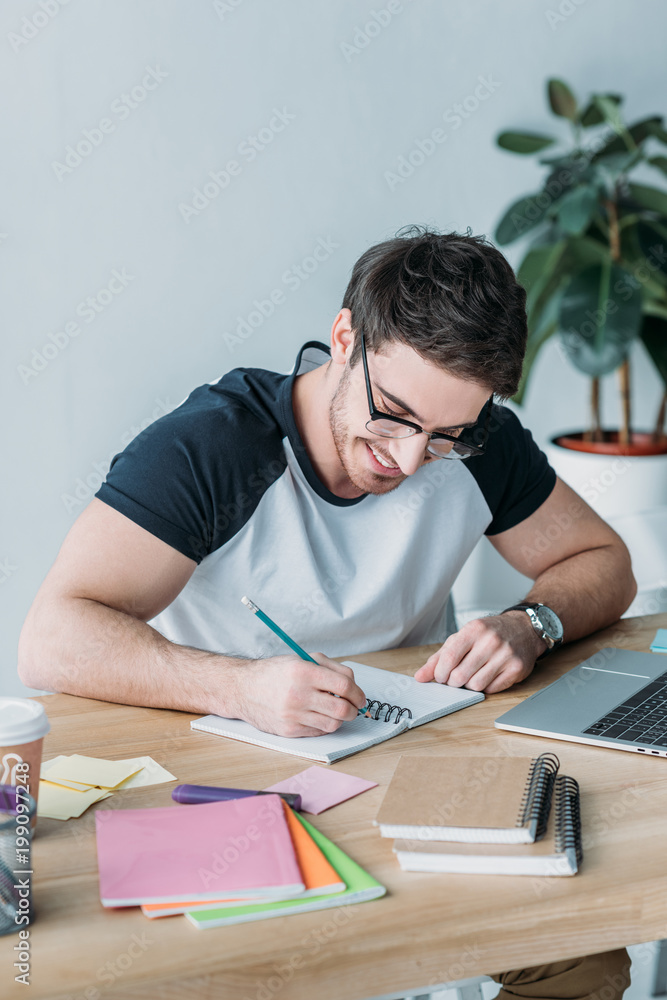 handsome smiling student in eyeglasses writing in copybook handsome smiling student in eyeglasses writing in copybook