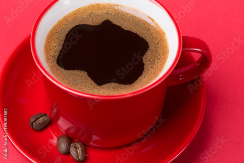 red cup with a drink of coffee with foam on a red background, near coffee beans on a saucer