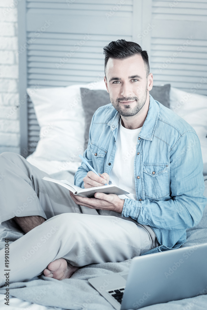 Fototapeta premium Positive mood. Delighted handsome positive man holding a notebook and looking at you while sitting on the bed