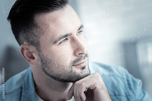 Thoughtful look. Nice handsome young man holing his chin and looking in front of him while being thoughtful