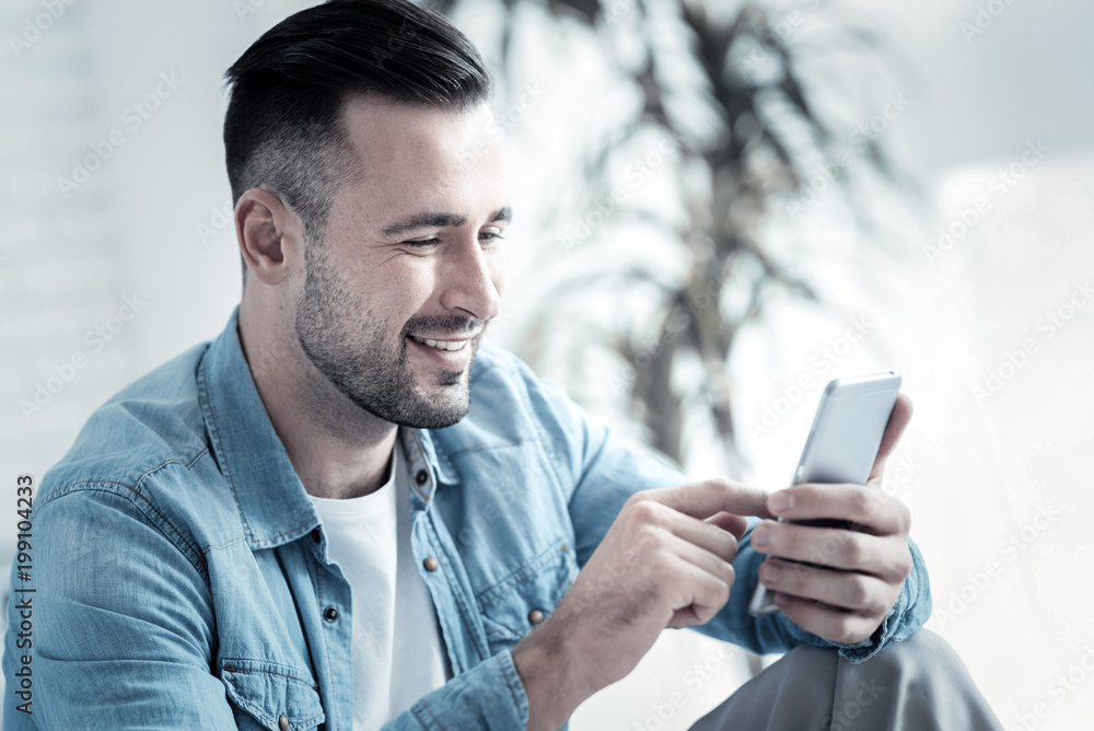Modern device. Delighted positive happy man sitting at home and using his smartphone while typing a message Modern device. Delighted positive happy man sitting at home and using his smartphone while typing a message