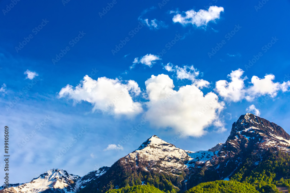 Surprisingly beautiful mountain range with melted snow and bright green forest at the foot with clean white clouds above them and a blue colorful sky. Caucasus mountains landscapes, Sochi, Russia.