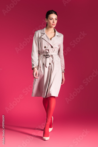 Woman walking in beige trench and heel shoes on red background