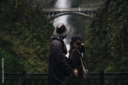 Obraz na plátně Hugging Asian couple stands before a beautiful waterfall in the mountains