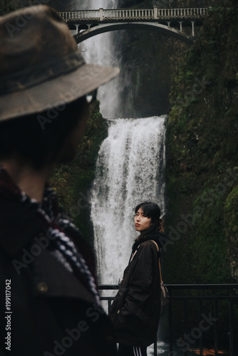 Man looks at an Asian woman standing before a waterfall somewhere in the mountains