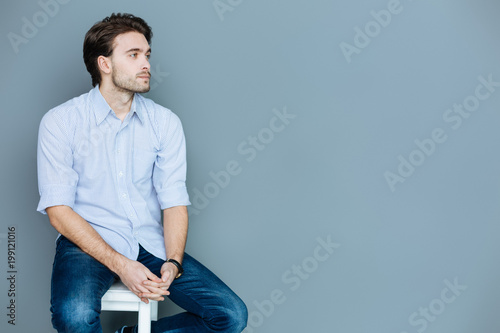 I am tired. Pleasant sad young man sitting on a stool and holding his hands together while thinking about his life