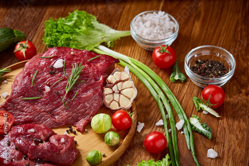 Still life of raw beef meat with vegetables on wooden plate over white background, top view, selective focus