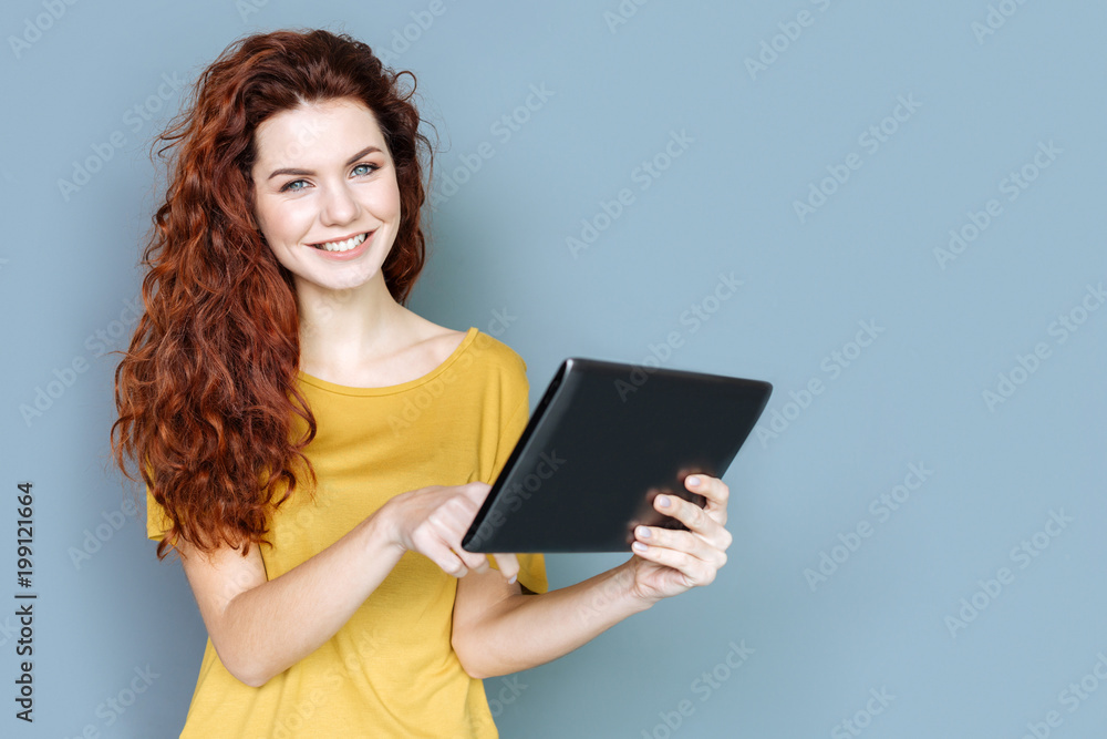 Digital technology. Attractive positive young woman smiling and looking at you while using a modern tablet Digital technology. Attractive positive young woman smiling and looking at you while using a modern tablet