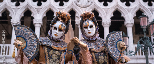Two women in traditional costume and painted masks, with decorated fans, standing in front ofthe Doges Palace during the Venice Carnival (Carnivale di Venezia) © Lois GoBe