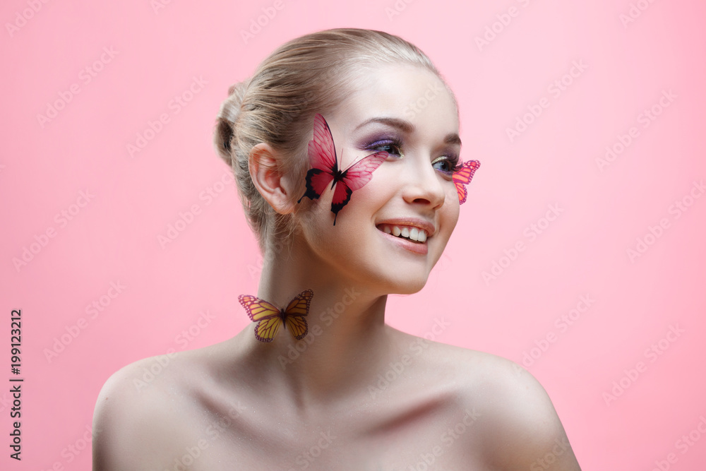 Beauty fashion portrait of a beautiful smiling girl with a gentle make-up and butterflies on her face isolated on a pink background.