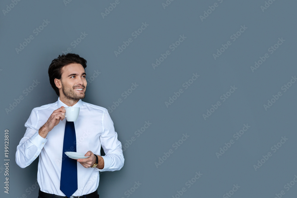 Coffee break. Joyful positive handsome man smiling and enjoying his coffee while having a break at work