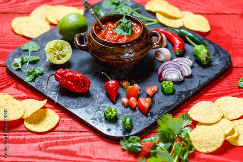 Homemade salsa with ingredients on black stone desk on red rustic background, tomato, pepper, onion, chili, habanero, coriander leaf. Mexican food concept, top view, overhead